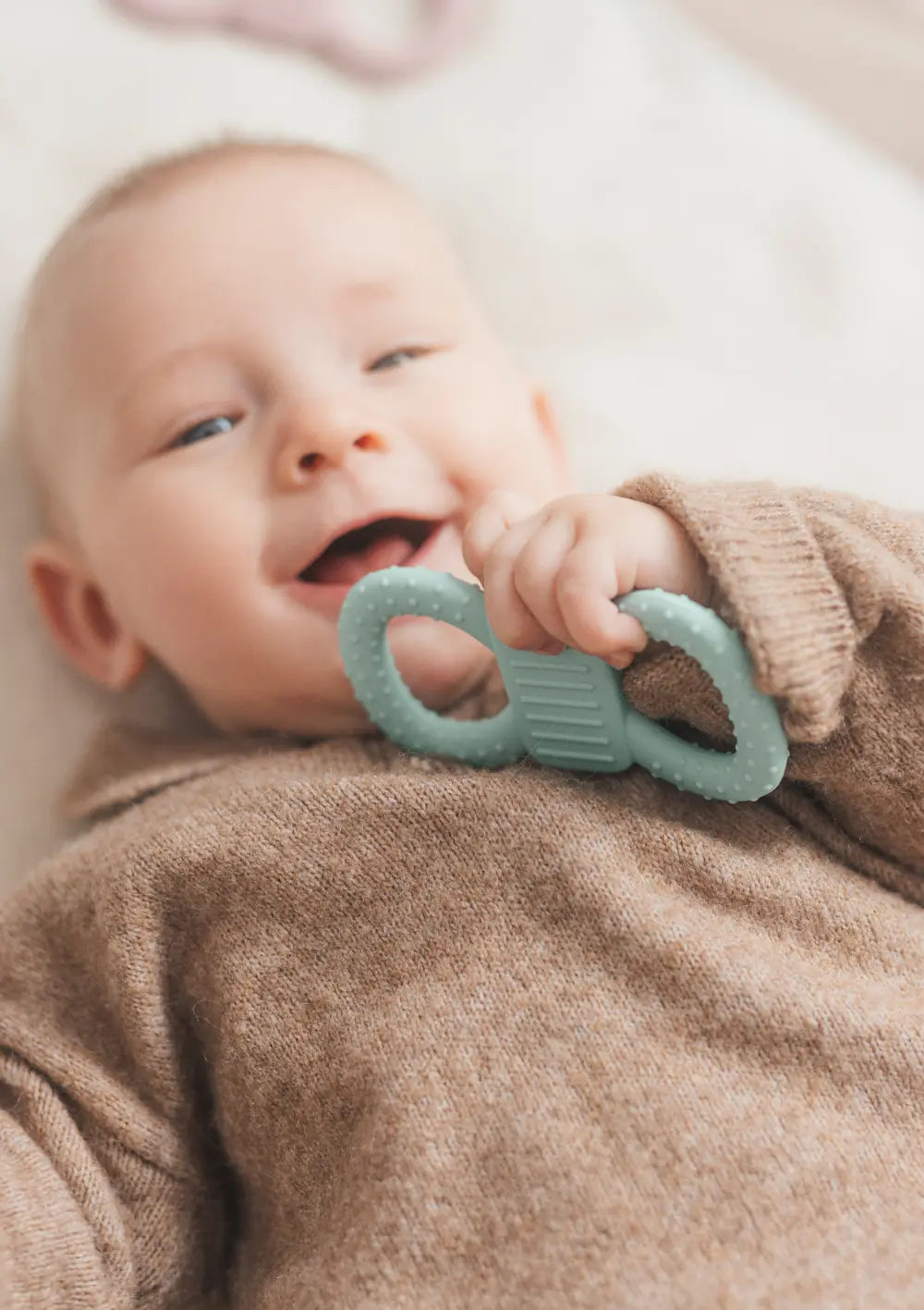 Lachendes Baby hält mimiloo Silikon Lern- und Beißring Mint beim Liegen auf einer hellen Decke.